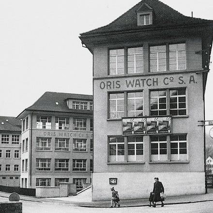 Two people walk past the Oris Watch Co. S.A. factory buildings in a black and white street scene.
