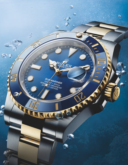 A Rolex Submariner watch with a blue and gold bezel is underwater, surrounded by bubbles.