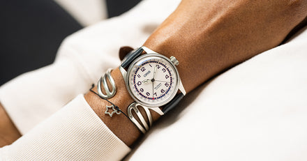 A wristwatch with a white dial and black strap on a person’s wrist, next to silver bracelets.