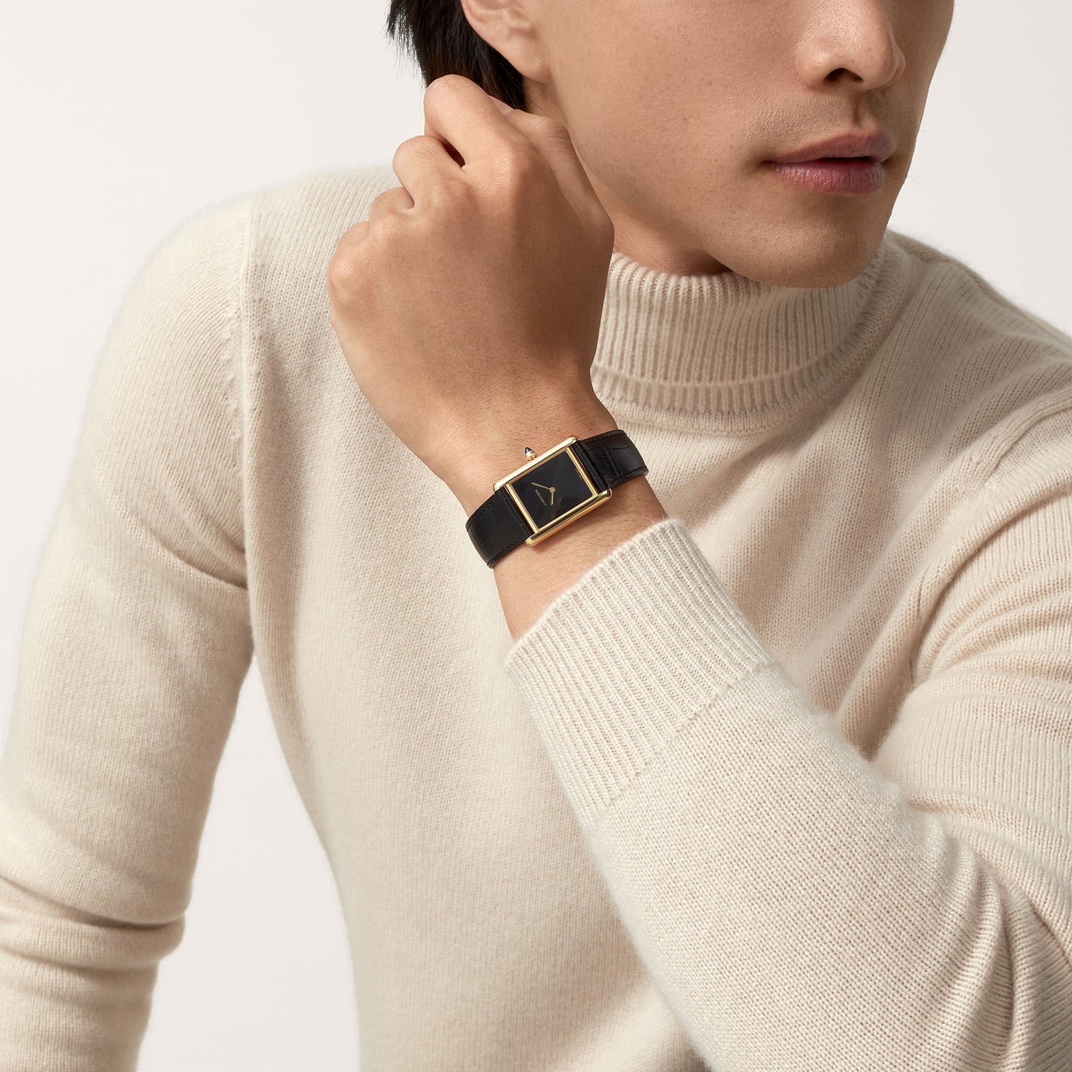 A person in a cream turtleneck displays a Cartier Tank Louis Cartier gold watch with a black strap on their wrist.