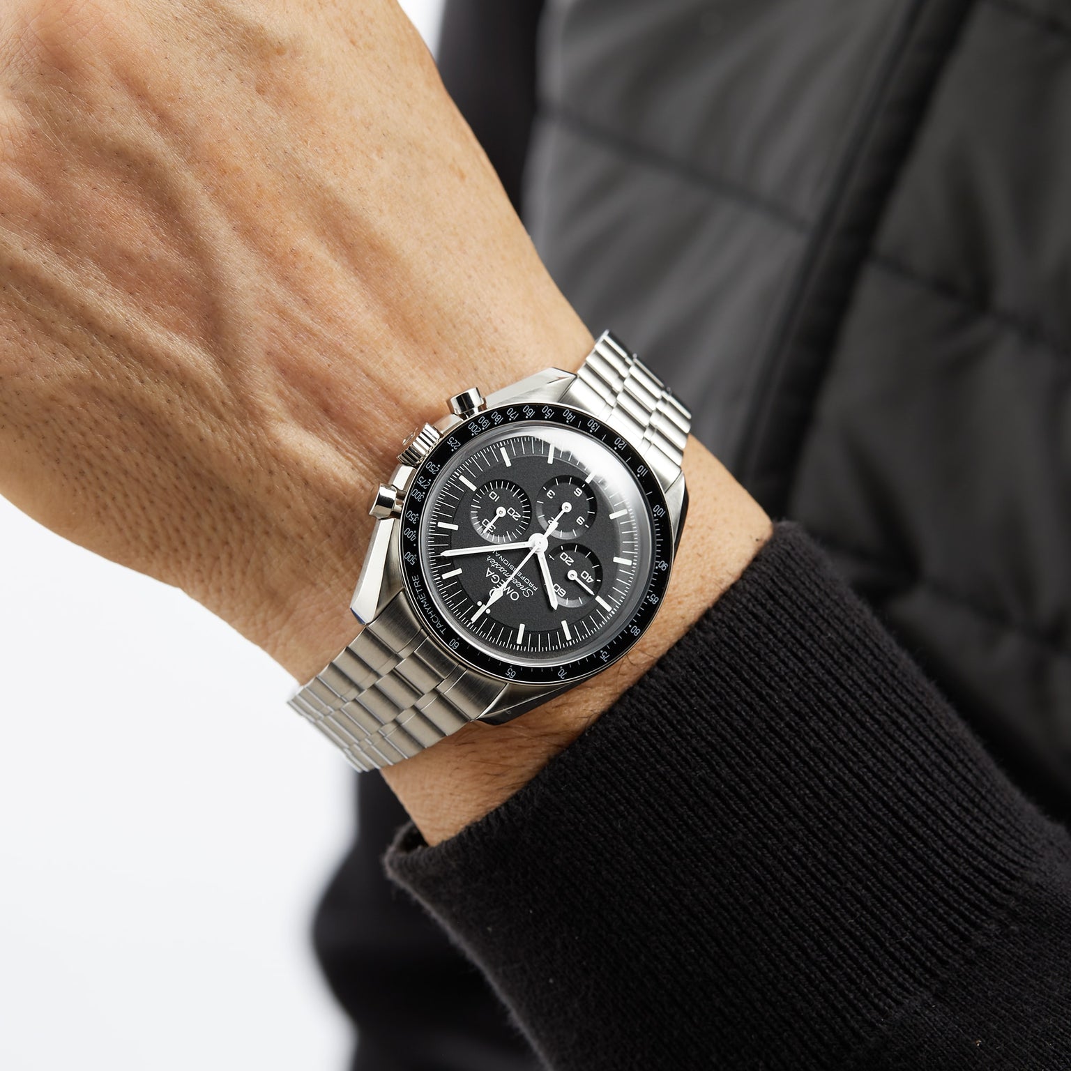 A person wears an OMEGA Speedmaster Moonwatch Professional with a black dial and three subdials, paired with a black quilted jacket.