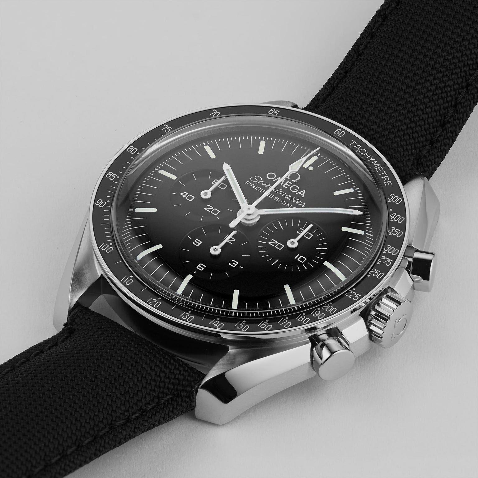 A close-up of the OMEGA Speedmaster Moonwatch Professional with a black dial, three subdials, black fabric strap, and precise 3861 calibre movement, photographed on a light background.