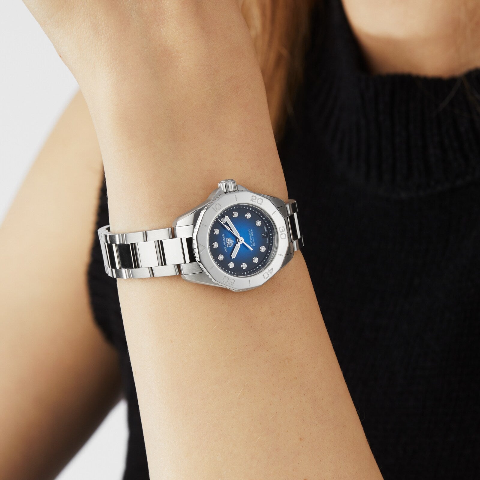 A person in a sleeveless black turtleneck wears the TAG Heuer Aquaracer Professional 200 Date, featuring a blue face, diamond markers, and a stainless steel case.