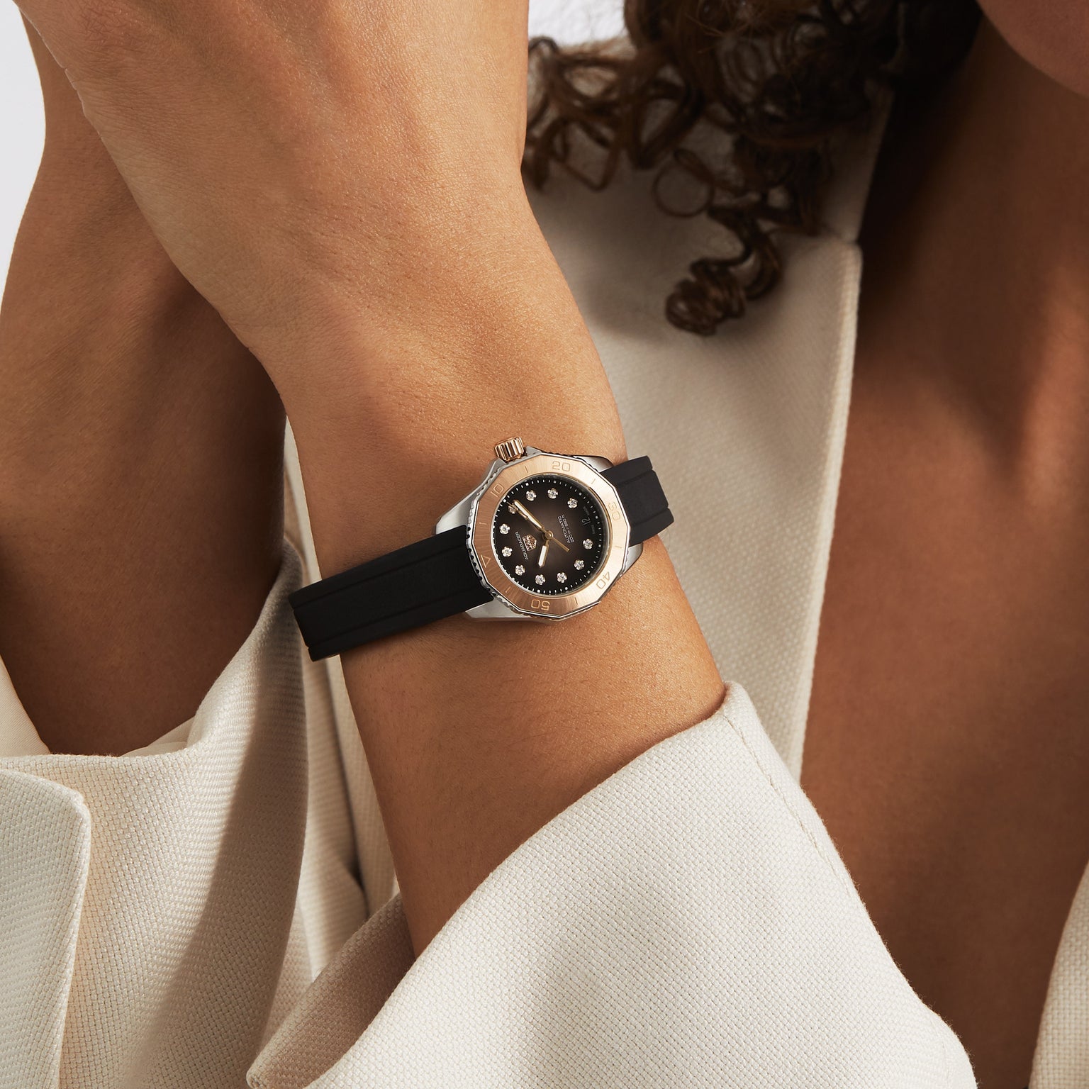 A person in a beige jacket rests their arm on their head, showcasing a TAG Heuer Aquaracer Professional 200 Date watch with a silver case, black strap, black dial, and notable water resistance.