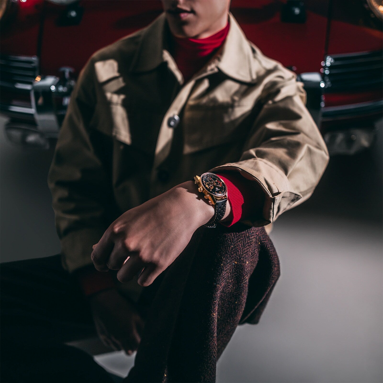 Wearing a beige jacket, a person showcases the TAG Heuer Carrera Chronosprint x Porsche Rallye wristwatch while seated in front of a red vintage car.