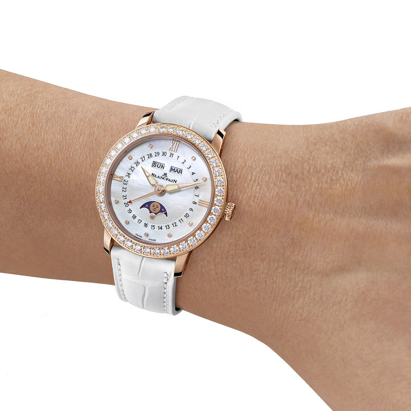 A wrist adorned with the Blancpain Ladybird Quantième Complet, featuring a white leather strap, diamond-set bezel, and an intricate moon phase dial showing date and day.