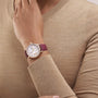 A person in a beige sweater rests their hand on their shoulder, showcasing the Vacheron Constantin Traditionnelle Moon Phase watch with pink gold, a burgundy leather strap, round white face, and an elegant moon phase detail.
