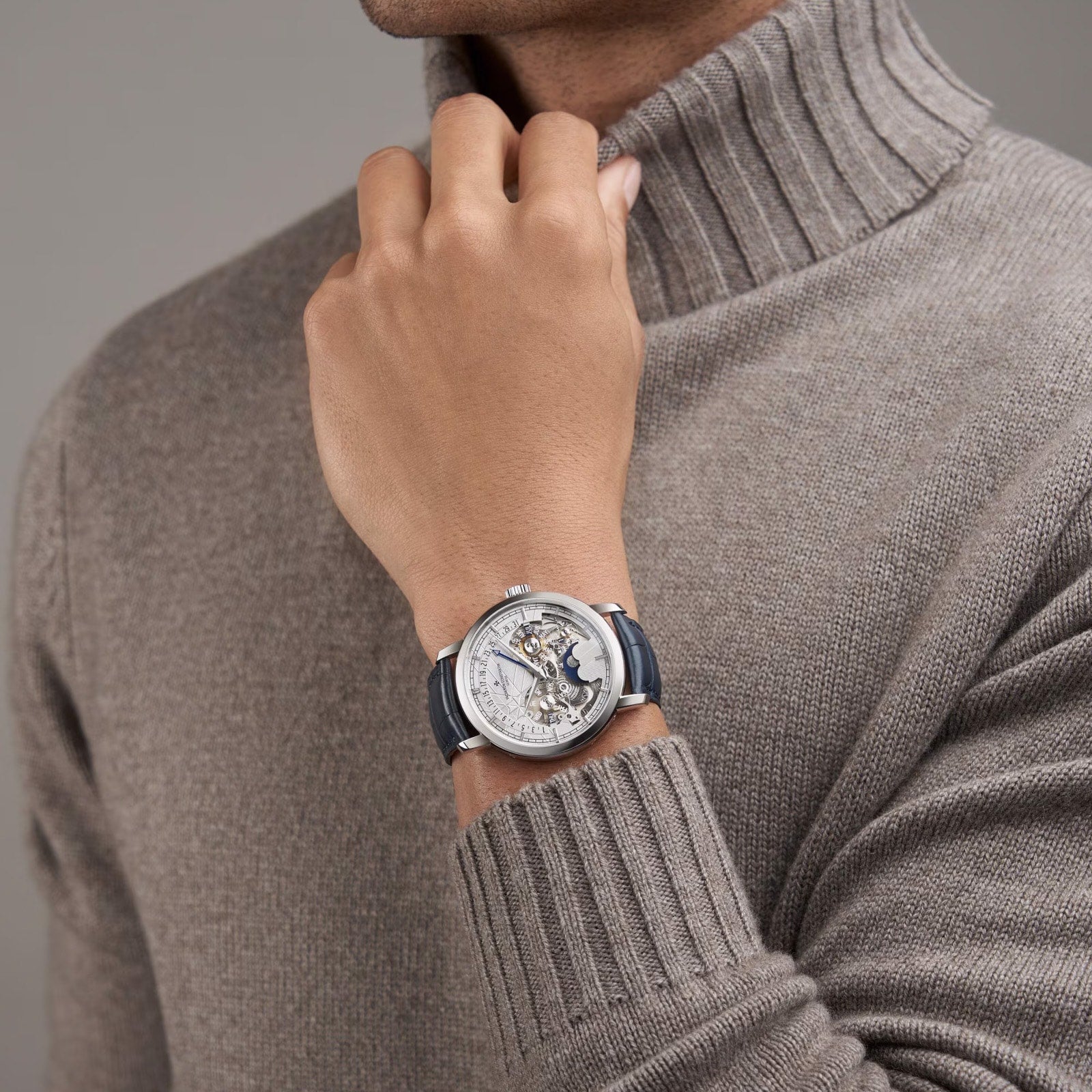 A person in a taupe ribbed turtleneck sweater showcases a Vacheron Constantin Traditionnelle Perpetual Calendar Retrograde Date Openface platinum watch with visible self-winding movement and a dark strap on their left wrist.