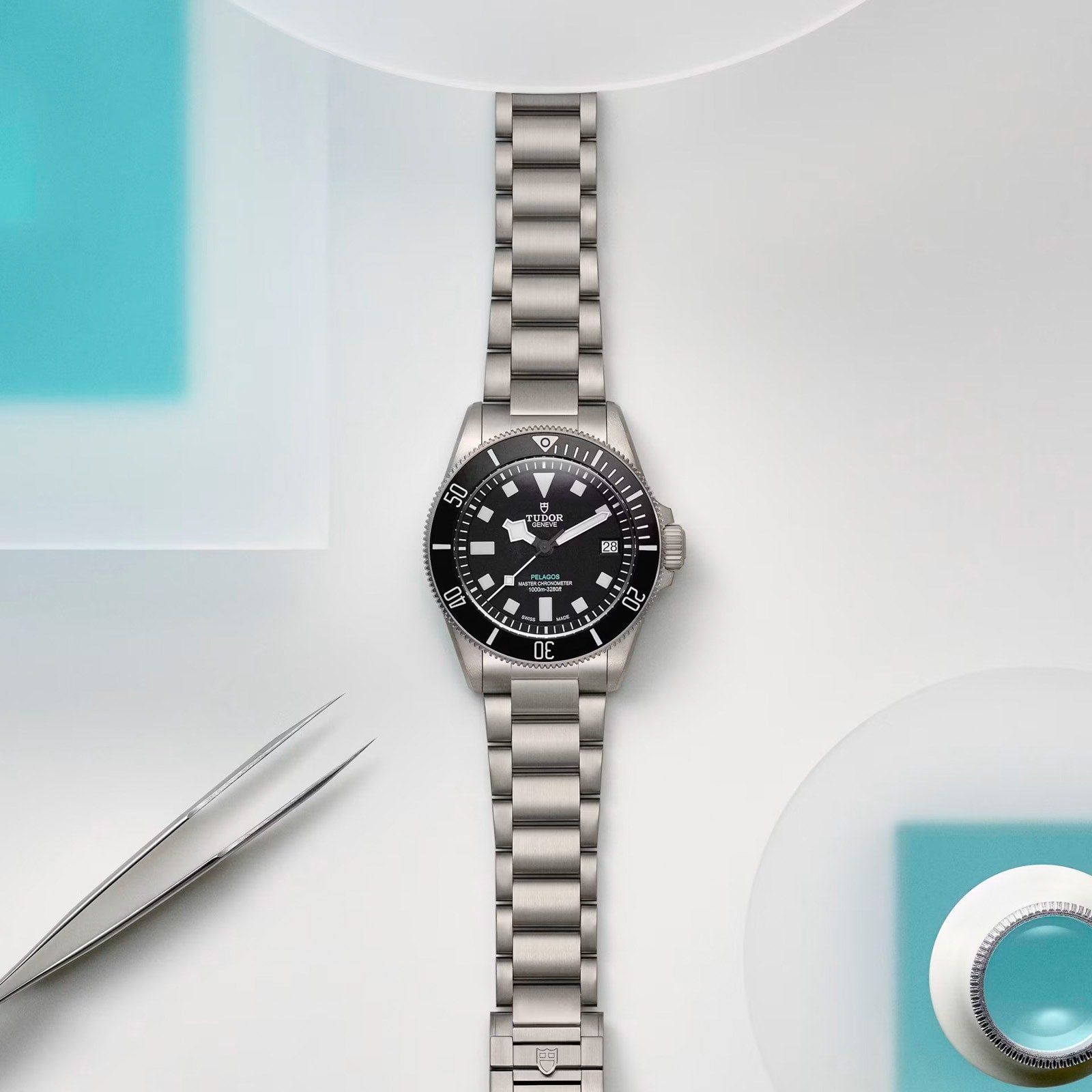 The TUDOR Pelagos Ultra, a silver wristwatch with a black dial on a metal bracelet, rests on a white surface featuring blue accents and nearby tweezers.