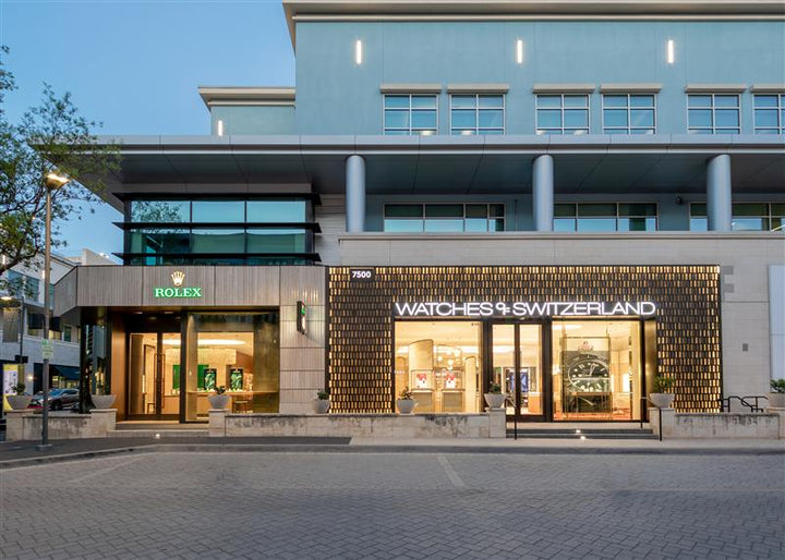 Street view of Rolex and Watches of Switzerland store fronts with large illuminated signs and modern architecture.