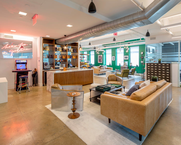 Modern office lounge with tan leather sofas, a bar area, arcade game, and green accent walls.