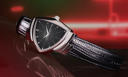 A Hamilton Ventura watch with a black leather strap and black dial sitting on a reflective red surface with a red background
