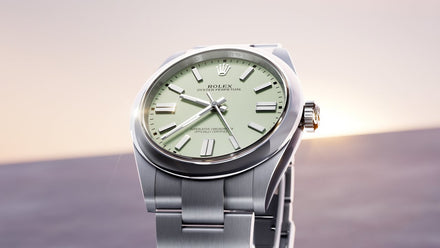 A close-up of a silver Rolex watch with a light green face, shown against a soft, blurred background.