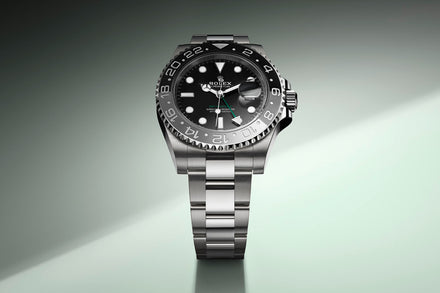 A stainless steel Rolex watch with a black dial and bezel, displayed on a gray-green gradient background.